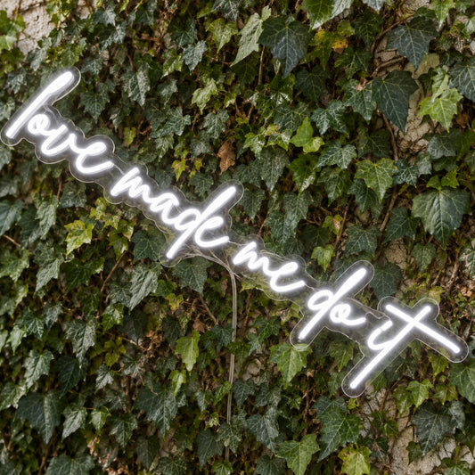 Love Made Me Do It LED Neon Light Sign in white, featuring script lettering against a green foliage wall, viewed at an angle.