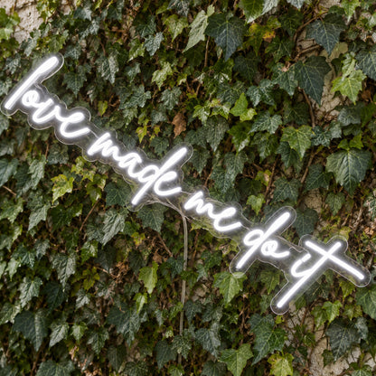 Love Made Me Do It LED Neon Light Sign in white, featuring script lettering against a green foliage wall, viewed at an angle.
