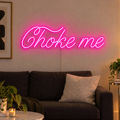 Choke Me Neon Sign in pink, featuring Choke Me script lettering above a dark sofa.