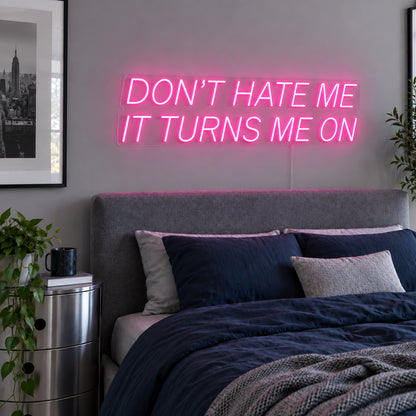 Don't Hate Me It Turns Me On Neon Sign glowing pink above a bed with dark blue bedding.