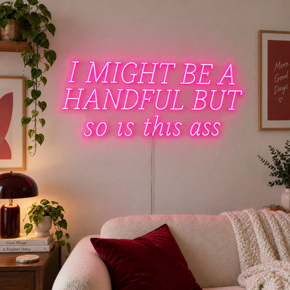 I Might Be A Handful But So Is This Ass Neon Sign glowing pink in wide lettering above a cream sofa.