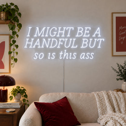 I Might Be A Handful But So Is This Ass Neon Sign glowing white in wide lettering above a cream sofa.