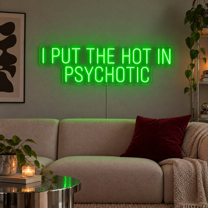 I Put The Hot In Psychotic Neon Sign with green lighting, displayed above a neutral sofa.
