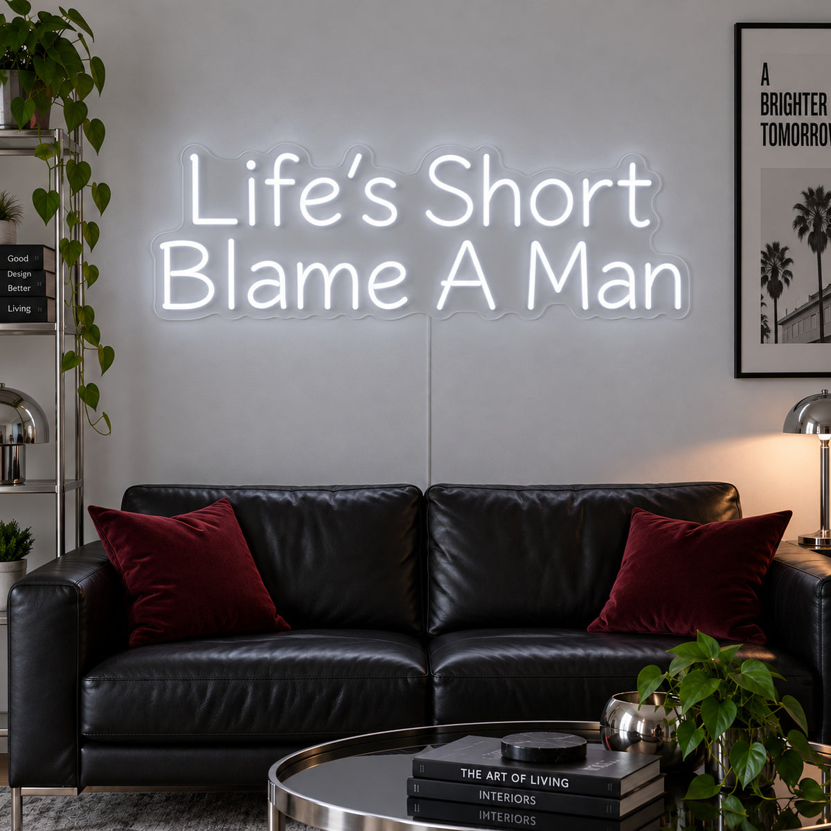 Life's Short Blame A Man Neon Sign glowing white above a black leather sofa.