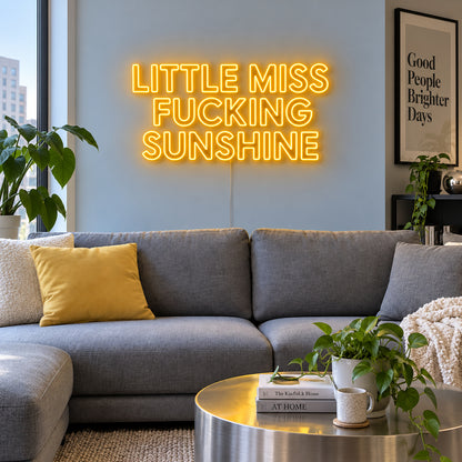Little Miss Fucking Sunshine Neon Sign glowing gold above a grey sofa with yellow cushions.