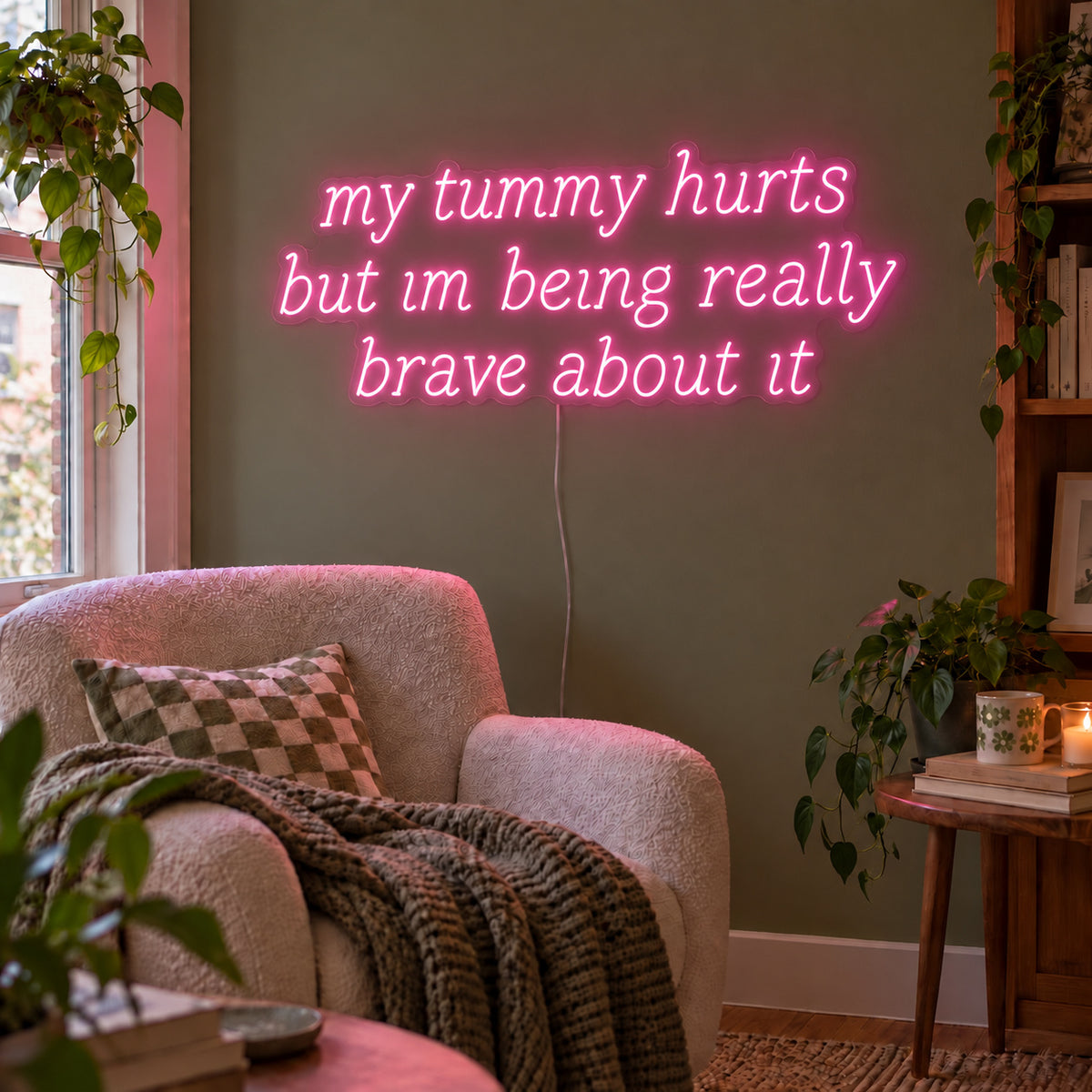 My Tummy Hurts But Im Being Really Brave About It Neon Sign glowing pink above a reading chair against a green wall.
