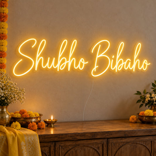 Shubho Bibaho Neon Sign on clear cut-to-shape acrylic, illuminated on a wall in a styled celebration setting