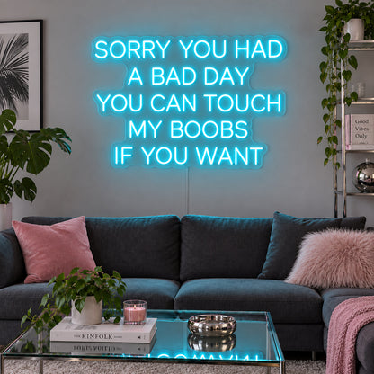Ice Blue version of the Sorry You Had A Bad Day You Can Touch My Boobs If You Want Neon Sign, shown above a grey sofa with pink cushions.